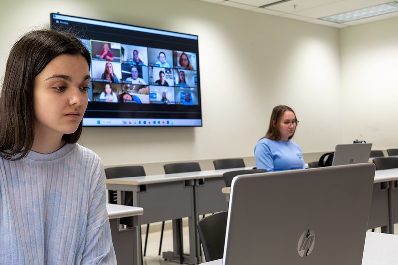 student sits in classroom with tv screen behind them. On the screen, multiple students are video calling into class